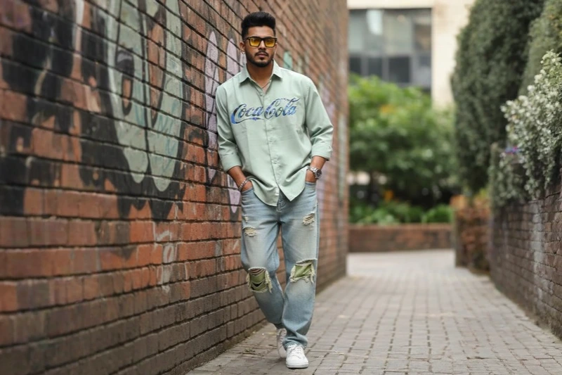 A man wearing a light green Coca-Cola shirt, ripped jeans, and white sneakers, leaning against a brick wall in an urban alley.